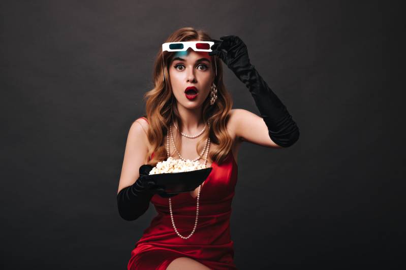 Surprised lady in red dress taking off 3D glasses and holding popcorn. Shocked woman in silk festive outfit watching movie on black background